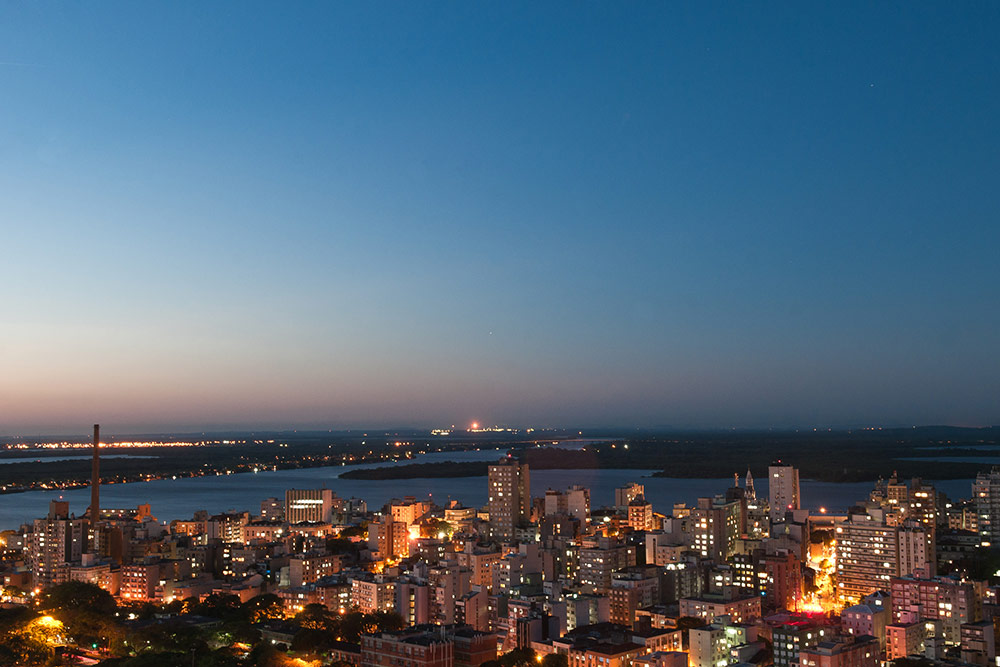 Vista aérea da cidade de Porto Alegre ao anoitecer. Prédios com luzes acesas no primeiro plano, tendo ao fundo o lago Guaíba abaixo de um céu azul escuro.